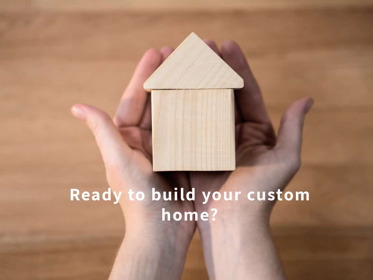 Pair of hands carefully holding two pieces of wood blocks forming a home. Text reads, "Ready to build your custom home?" Blurred background appears to be a wood panel, backdrop.