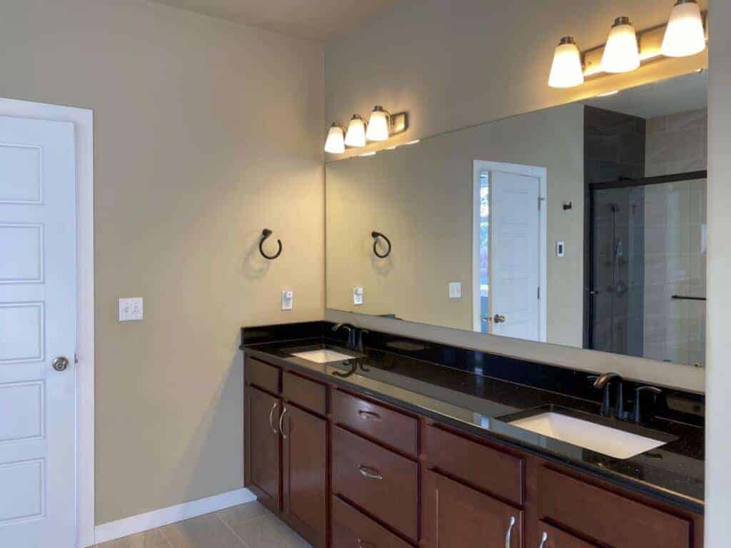 Two bathroom sinks, mirror, large walk-in shower, (Only viewed in the mirror) dark wood cabinetry with black countertops with large stone tile flooring, and a single bathroom door.