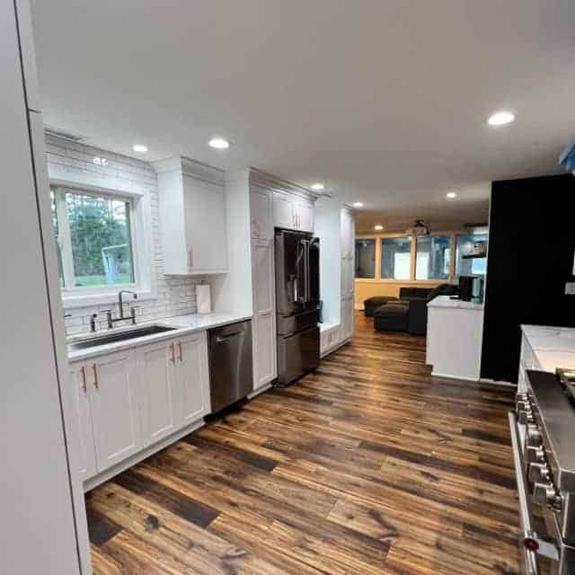 Full view of a remodeled kitchen, new kitchen appliances, and hardwood laminate flooring. Kitchen sink, and faucet with another living area in the background.